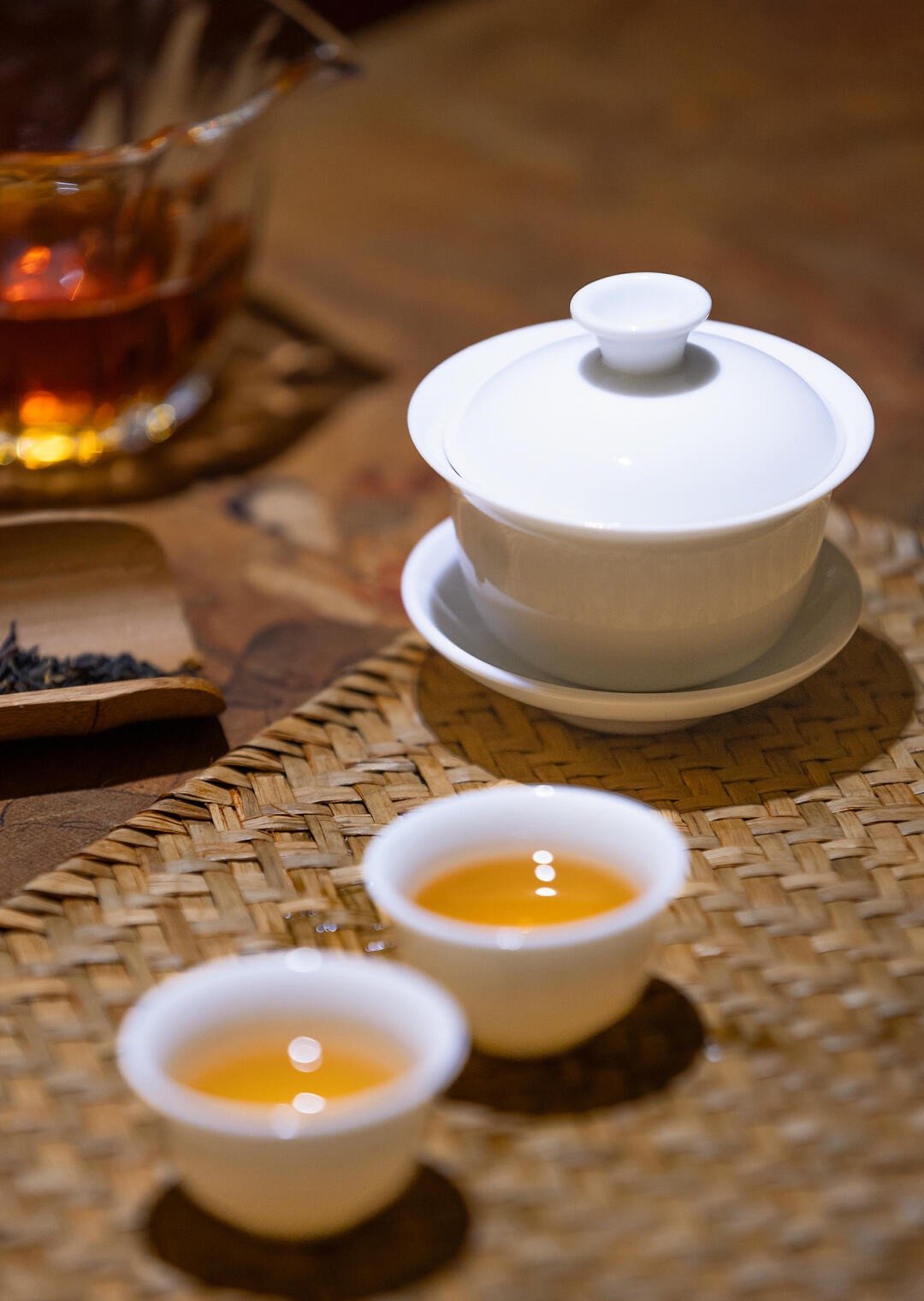 The Origins of Tea White porcelain gaiwan with two small tea cups filled with golden oolong tea on a woven bamboo mat